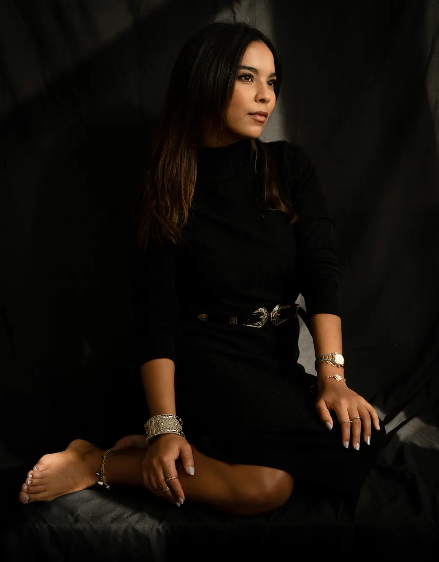 Mujer con vestido negro y joyería de oro sentada en un estudio